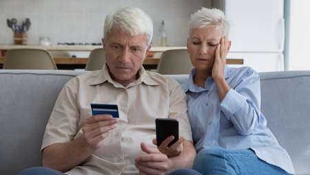 Photo of two senior citizens looking at a cell phone and holding thier credit card. They are visibly upset.